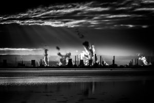 Ciudad contaminada en blanco y negro con nubes y chimeneas de fábricas al fondo y un mar calmado en primer plano